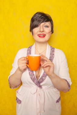 kısa saçlı sevimli kadın rahat pembe bornoz giymiş ve sarı sağlam bir arka plan stüdyo üzerinde turuncu bir fincan çay içer