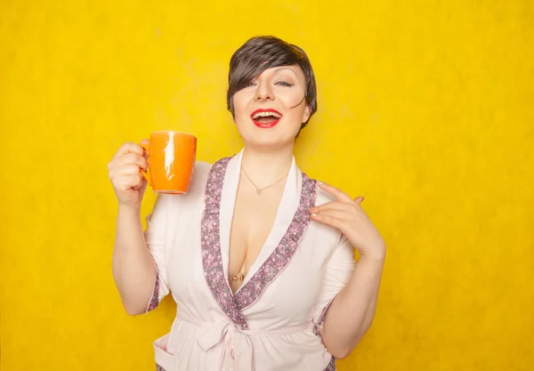 短い髪のかわいい女性が居心地の良いピンクのバスローブに身を包んだし スタジオで黄色の固体背景にオレンジ色のカップからお茶を飲む — ストック写真