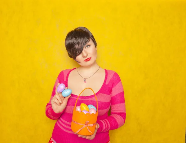 漂亮的成人胖乎乎的女人短发举行五颜六色的节日鸡蛋和复活节篮子上黄色固体工作室的背景 — 图库照片