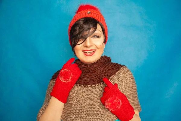 pretty brunette girl wearing fashion red knitted winter gat and warm beautiful gloves on blue studio solid background