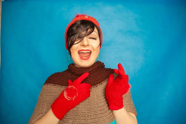 pretty brunette girl wearing fashion red knitted winter gat and warm beautiful gloves on blue studio solid background