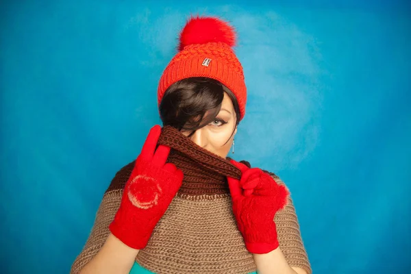 pretty brunette girl wearing fashion red knitted winter gat and warm beautiful gloves on blue studio solid background