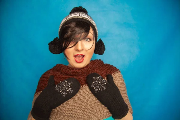 pretty brunette girl wearing fashion black knitted winter hat and warm beautiful mittens on blue studio solid background