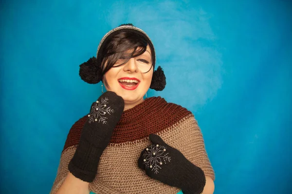 pretty brunette girl wearing fashion black knitted winter hat and warm beautiful mittens on blue studio solid background