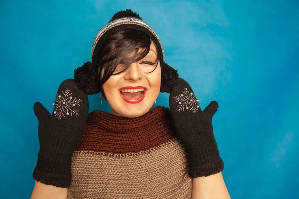 pretty brunette girl wearing fashion black knitted winter hat and warm beautiful mittens on blue studio solid background