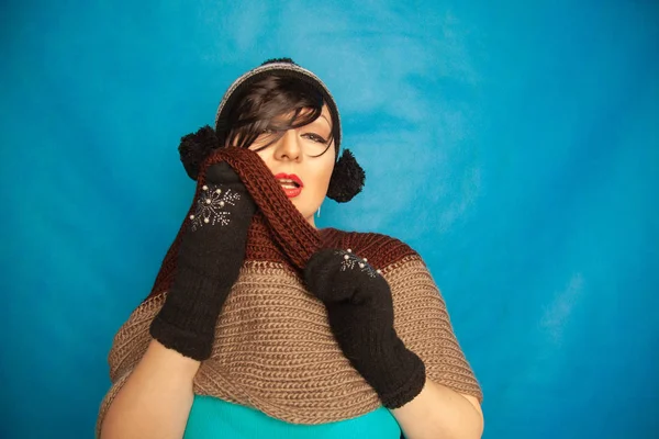 pretty brunette girl wearing fashion black knitted winter hat and warm beautiful mittens on blue studio solid background