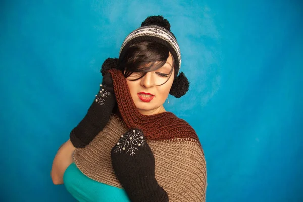 pretty brunette girl wearing fashion black knitted winter hat and warm beautiful mittens on blue studio solid background