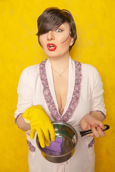 バスローブ姿で美しい明るいブルネットは 鍋と食器洗いスポンジを保持します イエロー スタジオの背景に若い女性主婦 — ストック写真