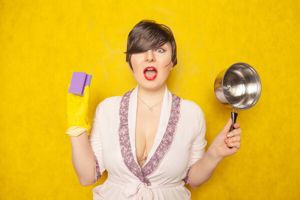beautiful bright brunette in a Bathrobe holds a pan and a sponge for washing dishes. young woman housewife on yellow studio background. 