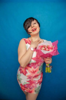 Bahar elbise sevimli gülümseyen kız stüdyoda mavi bir arka plan üzerinde tulip buket pembe taze tutar. o gerçekten hediye ve kesinlikle mutlu bulunuyor.