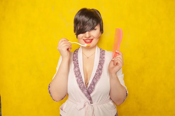 cute chubby girl in a pink robe stands with a comb and toothbrush on a yellow background in the Studio