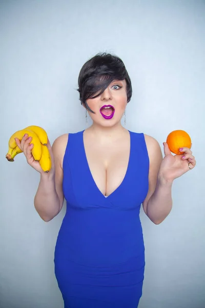 cute chubby girl with short black hair in a blue dress holding an orange and bananas in her hands, smiling with love for fruits on a white solid background in the Studio