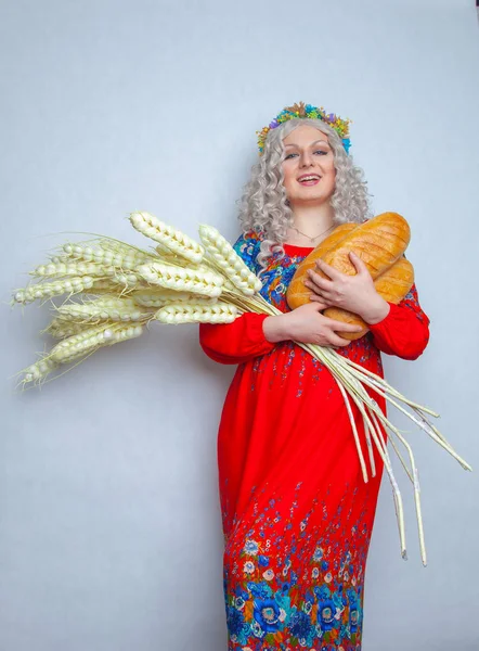 charming chubby smiling girl with big ears of ripe wheat and fresh bread in her hands. woman dressed in a traditional rustic costume and stands on white solid background in the Studio.