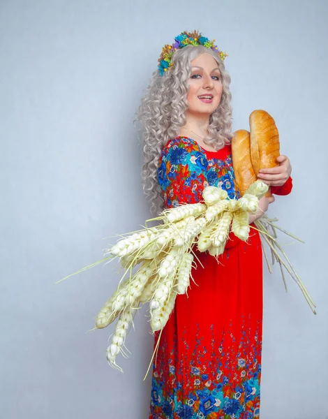 charming chubby smiling girl with big ears of ripe wheat and fresh bread in her hands. woman dressed in a traditional rustic costume and stands on white solid background in the Studio.