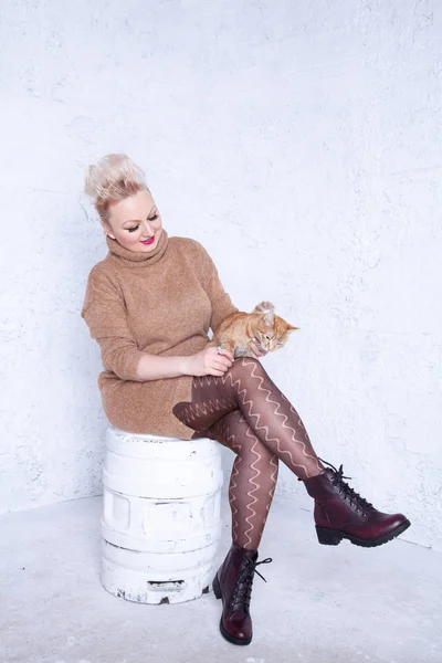 pretty plump young woman with short hair wearing brown sweater and having tenderness with her redhaired cat friend on white studio background