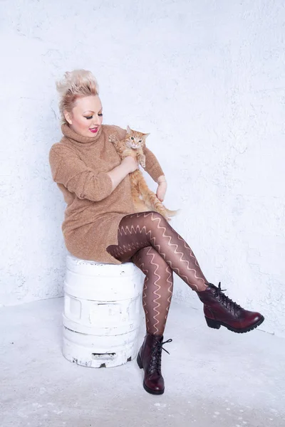 pretty plump young woman with short hair wearing brown sweater and having tenderness with her redhaired cat friend on white studio background