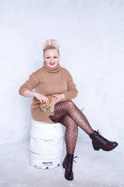 pretty plump young woman with short hair wearing brown sweater and having tenderness with her redhaired cat friend on white studio background