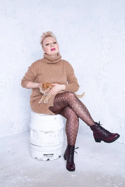 pretty plump young woman with short hair wearing brown sweater and having tenderness with her redhaired cat friend on white studio background