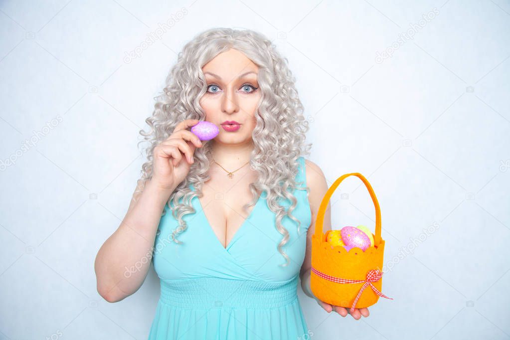 charming smiling blonde teenager stands in a blue dress with an orange basket with painted eggs for Easter alone on white studio solid background