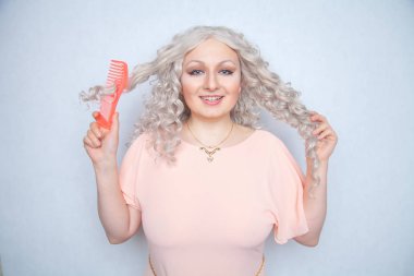 girl with curly white hair can't comb her loose dry hair and is upset about it. a blonde woman stands in a pink dress with an orange comb on a white Studio background. clipart