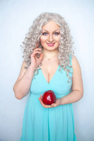 blonde curly girl in blue dress holding tasty big red apple and smile on white studio solid background