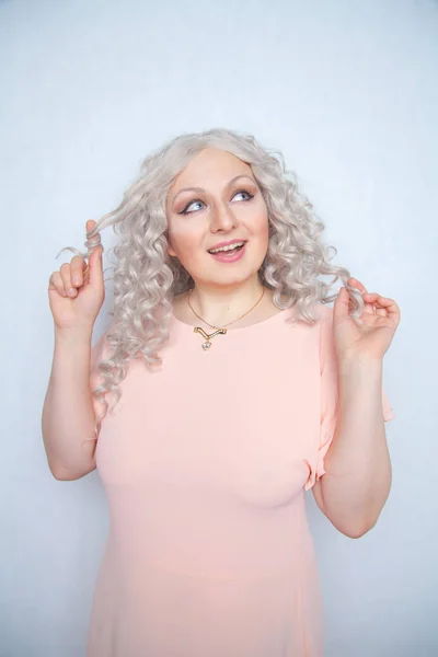 carefree girl in a pink dress twists her blonde curly hair on her finger and rejoices on a white monochrome Studio background