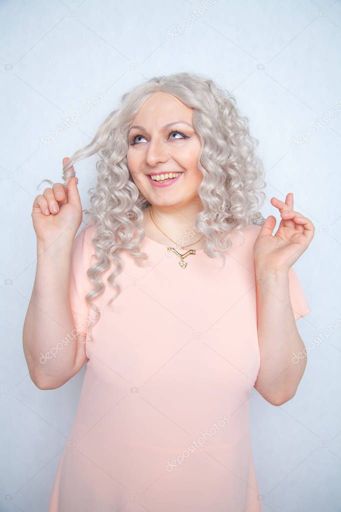 carefree girl in a pink dress twists her blonde curly hair on her finger and rejoices on a white monochrome Studio background