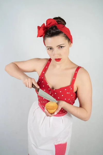 dangerous pin up girl housewife in red vintage polka dot dress stands with a huge kitchen knife in her hands and angrily rejoices on white solid studio background