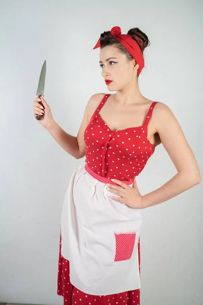 危险的别针女孩家庭主妇在红色的老式波尔卡点礼服站在她的手一个巨大的菜刀 愤怒地高兴在白色坚实的工作室背景 — 图库照片