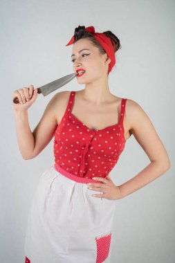 Kırmızı puantiyeli pinup elbise ve önlük duran, büyük bir bıçak tutarak ve beyaz stüdyo düz bir arka plan üzerinde tek başına yemek sıkışmış çünkü dişlerini malzeme çekme güzel genç kız