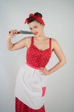 Kırmızı puantiyeli pinup elbise ve önlük duran, büyük bir bıçak tutarak ve beyaz stüdyo düz bir arka plan üzerinde tek başına yemek sıkışmış çünkü dişlerini malzeme çekme güzel genç kız