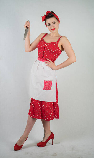 dangerous pin up girl housewife in red vintage polka dot dress stands with a huge kitchen knife in her hands and angrily rejoices on white solid studio background