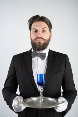 elini ve bir kadeh şarap beyaz sağlam bir arka plan stüdyo üzerinde çelik bir tepsi ile resmi iş takım elbiseli sakallı beyaz hizmetçi duruyor