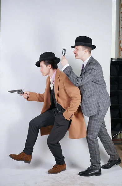 two caucasian detective men in retro suits with vintage hats sneak quietly go, holding a magnifying glass and a gun on white studio background