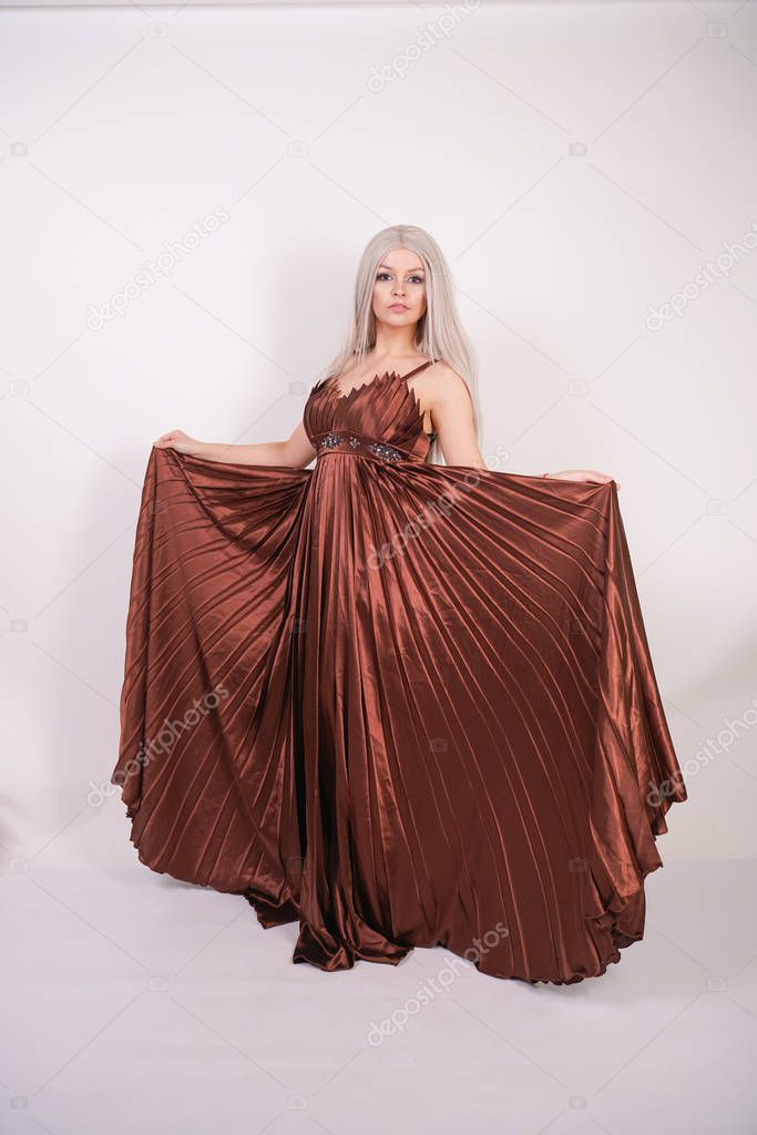 luxury blonde caucasian model girl in chocolate color long evening dress made of pleated fabric waving a flying dress and stands on a white Studio background