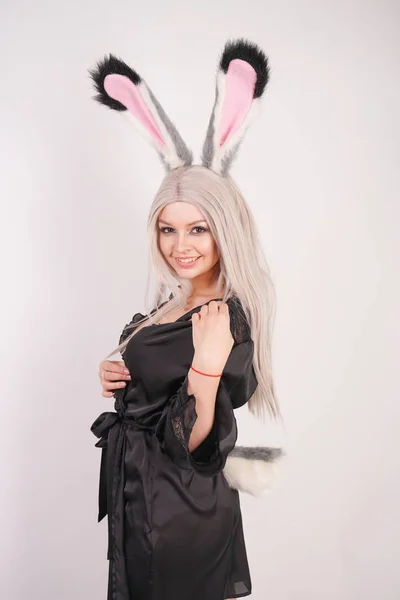 cute blonde fashion model posing in erotic silk black robe barefoot with big rabbit ears made of fur and a cute round tail on a white background in the Studio