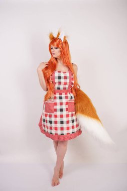 Tüylü büyük kulakları ile turuncu uzun peruk ve uzun kabarık kuyruk kırmızı mutfak ekose apron giyen beyaz bir arka planda yalnız yalınayak poz büyüleyici Fox kız