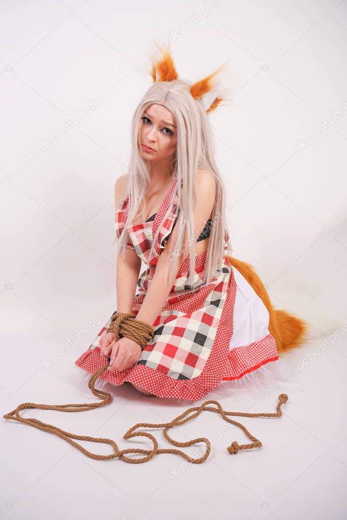 charming frightened blonde girl in a Fox costume with fur ears and a fluffy orange tail sits on her knees with her hands tied with a hemp rope on a white background in the Studio