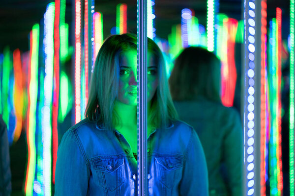 sweet caucasian girl walks in a mirror maze with colorful diodes and enjoys an unusual attraction room in the city