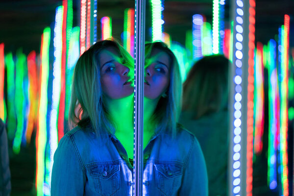 sweet caucasian girl walks in a mirror maze with colorful diodes and enjoys an unusual attraction room in the city