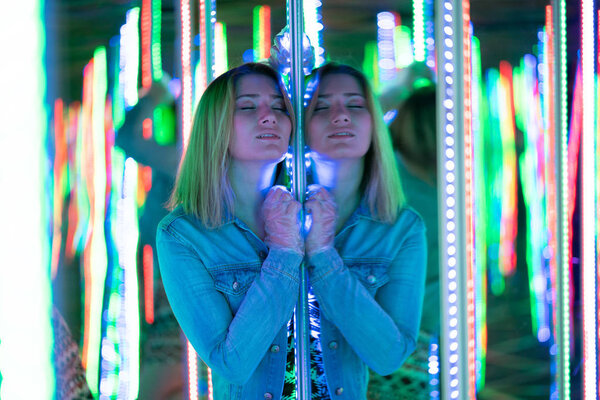 sweet caucasian girl walks in a mirror maze with colorful diodes and enjoys an unusual attraction room in the city