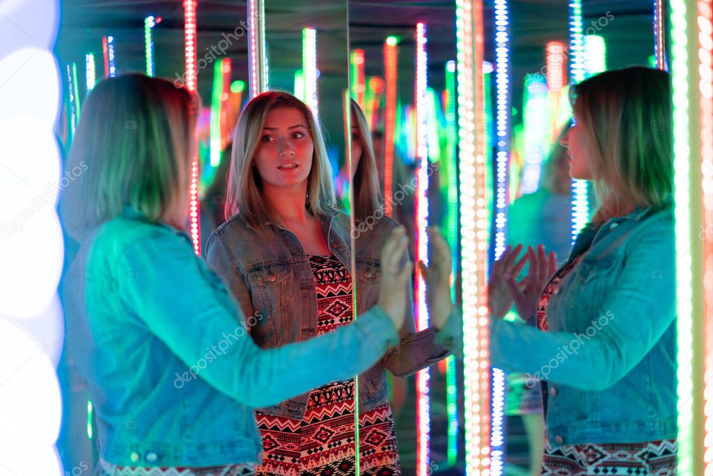 sweet caucasian girl walks in a mirror maze with colorful diodes and enjoys an unusual attraction room in the city