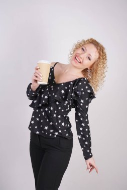 sevimli Kıvırcık sarışın kız siyah moda giysiler duruyor ve beyaz bir stüdyo arka planda bir kağıt fincan kahve tutar