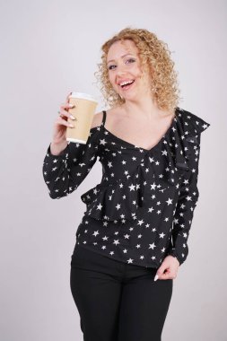 sevimli Kıvırcık sarışın kız siyah moda giysiler duruyor ve beyaz bir stüdyo arka planda bir kağıt fincan kahve tutar