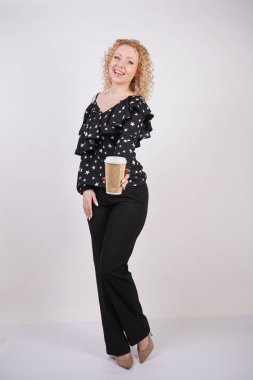 sevimli Kıvırcık sarışın kız siyah moda giysiler duruyor ve beyaz bir stüdyo arka planda bir kağıt fincan kahve tutar