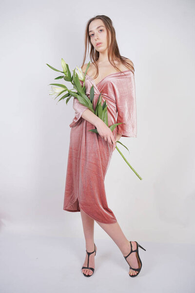shy caucasian girl with big eyes stands in a velvet pink dress and holds a white Lily in her hand as a symbol of innocence and purity. young woman posing on white studio background.