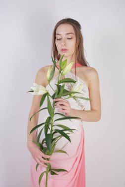 büyük gözleri ve kahverengi saçlı büyüleyici bir genç kız stüdyo beyaz bir arka planda bir moda elbise makyaj olmadan duruyor. Beyaz yemyeşil lilyum ile bir kök tutan güzel genç genç kadın.