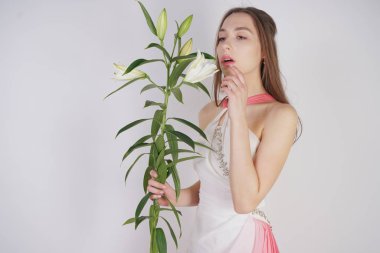 büyük gözleri ve kahverengi saçlı büyüleyici bir genç kız stüdyo beyaz bir arka planda bir moda elbise makyaj olmadan duruyor. Beyaz yemyeşil lilyum ile bir kök tutan güzel genç genç kadın.