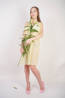 bir retro Sarı pilili şeffaf gecelik büyüleyici bir genç beyaz kız elleriyle yemyeşil bir zambak çiçek tutar ve stüdyo beyaz bir arka planda duruyor