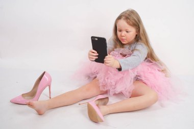 biraz beyaz çocuk kız oturur ve anne yüksek topuklu pembe ayakkabı koyar, beyaz stüdyo arka plan üzerinde onun akıllı telefonda tüm resimleri ve selfie 'ler alarak